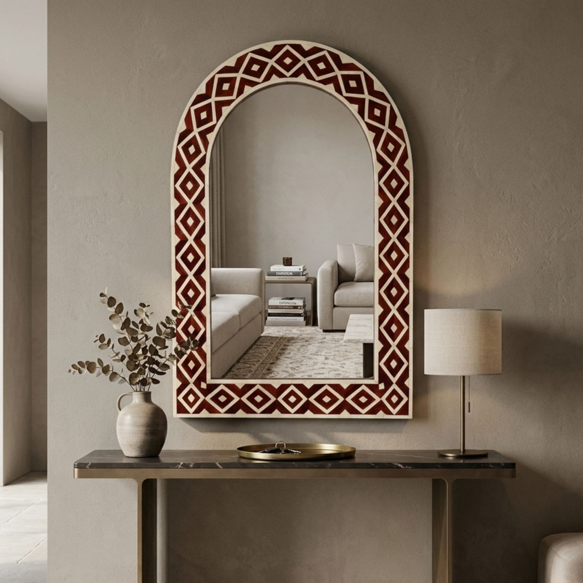 An arched decorative wall mirror with a geometric diamond-patterned frame in dark red and cream, styled on a console table in a neutral-toned living room setting.