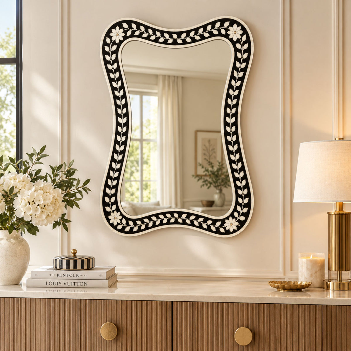 An ornate wavy rectangular wall mirror with a black and white floral bone-inlay frame, styled on a cream wall above a wooden console table with books, a lamp, and a vase of flowers.