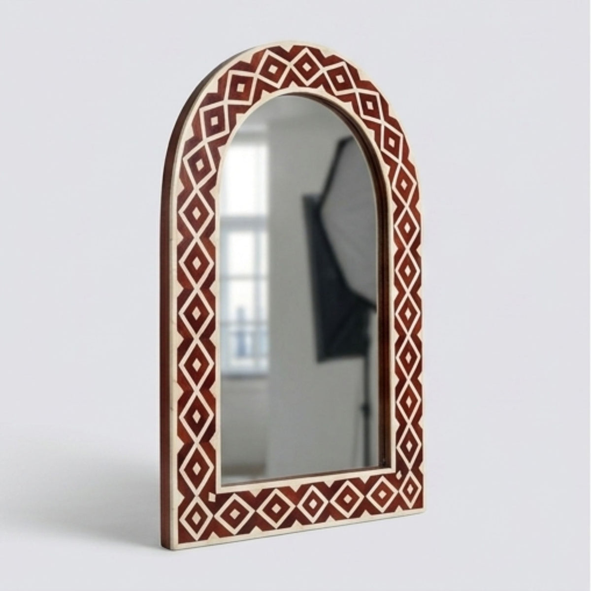 An arched wall mirror with a decorative border featuring a geometric diamond pattern in reddish-brown and cream, set against a light gray background.