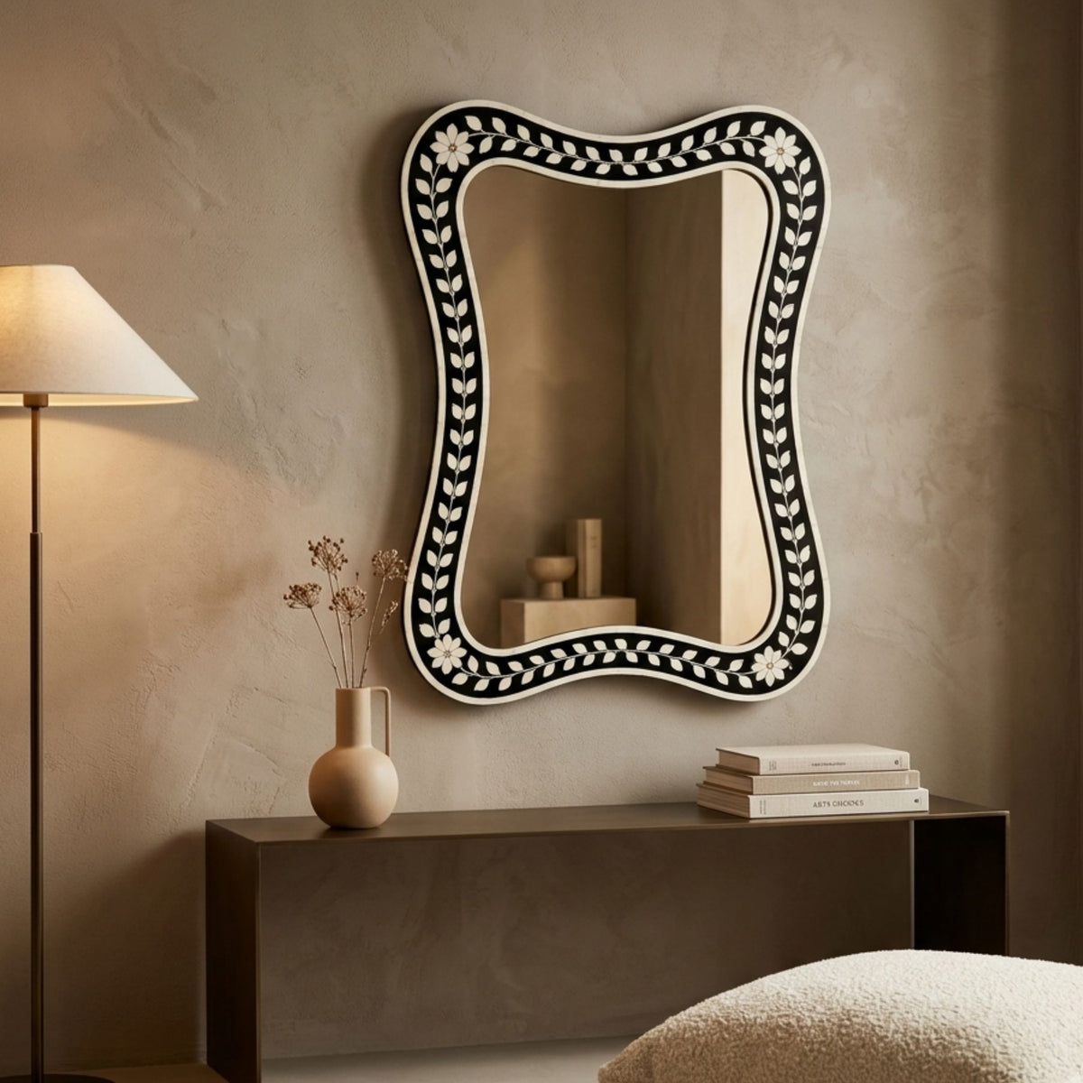 An asymmetrical bohemian-style wall mirror with a black and white floral inlay border, mounted on a textured beige wall above a dark console table with a lamp and books.