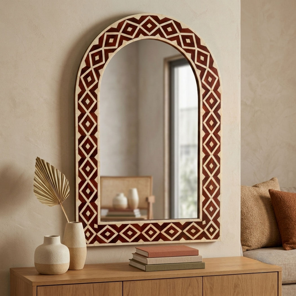 An arched wall mirror with a geometric diamond-patterned frame in dark reddish-brown and cream, styled on a light wood console table in a warm, neutral-toned room.