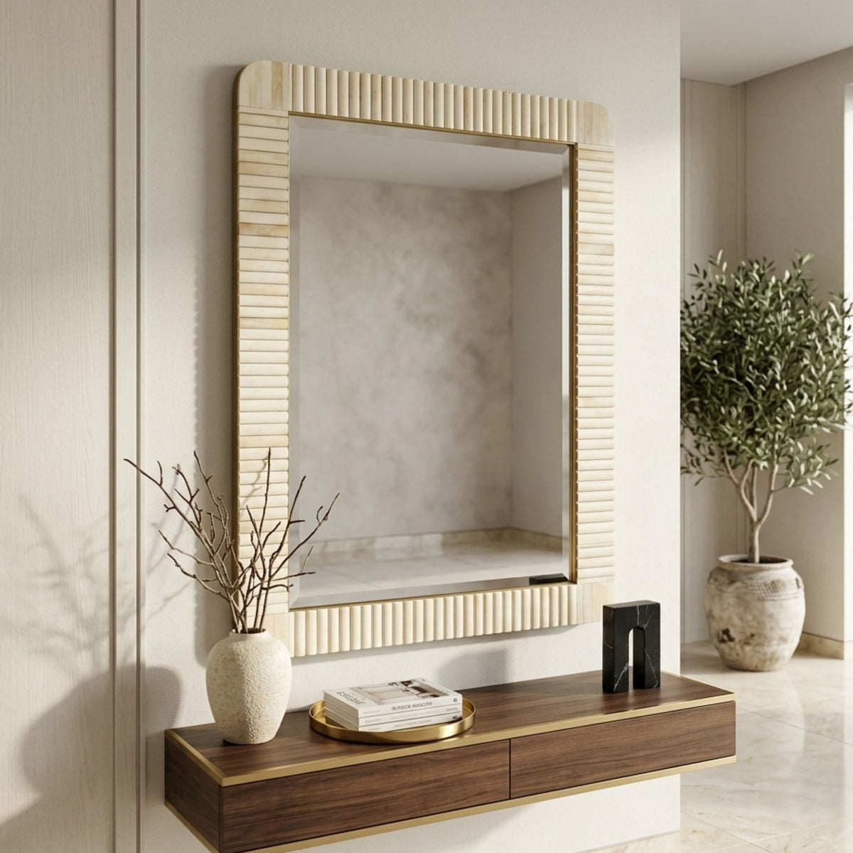 A large rectangular wall mirror with a fluted cream-beige frame and gold trim, mounted on a beige wall above a floating dark wood console table with gold accents. The scene is styled with a textured ceramic vase, a small stack of books, and a potted olive tree in a bright, modern interior.