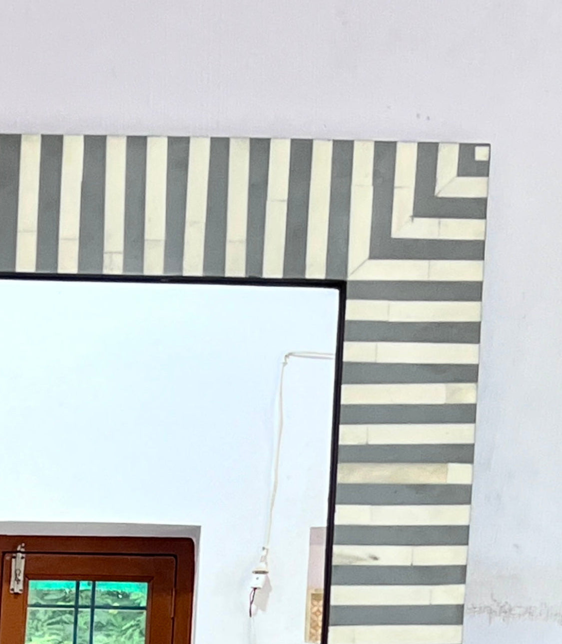 A large rectangular mirror with a black frame, set against a white wall with a decorative border of vertical and geometric striped tiles. A wooden door with glass panes is visible in the lower-left corner of the mirror reflection.