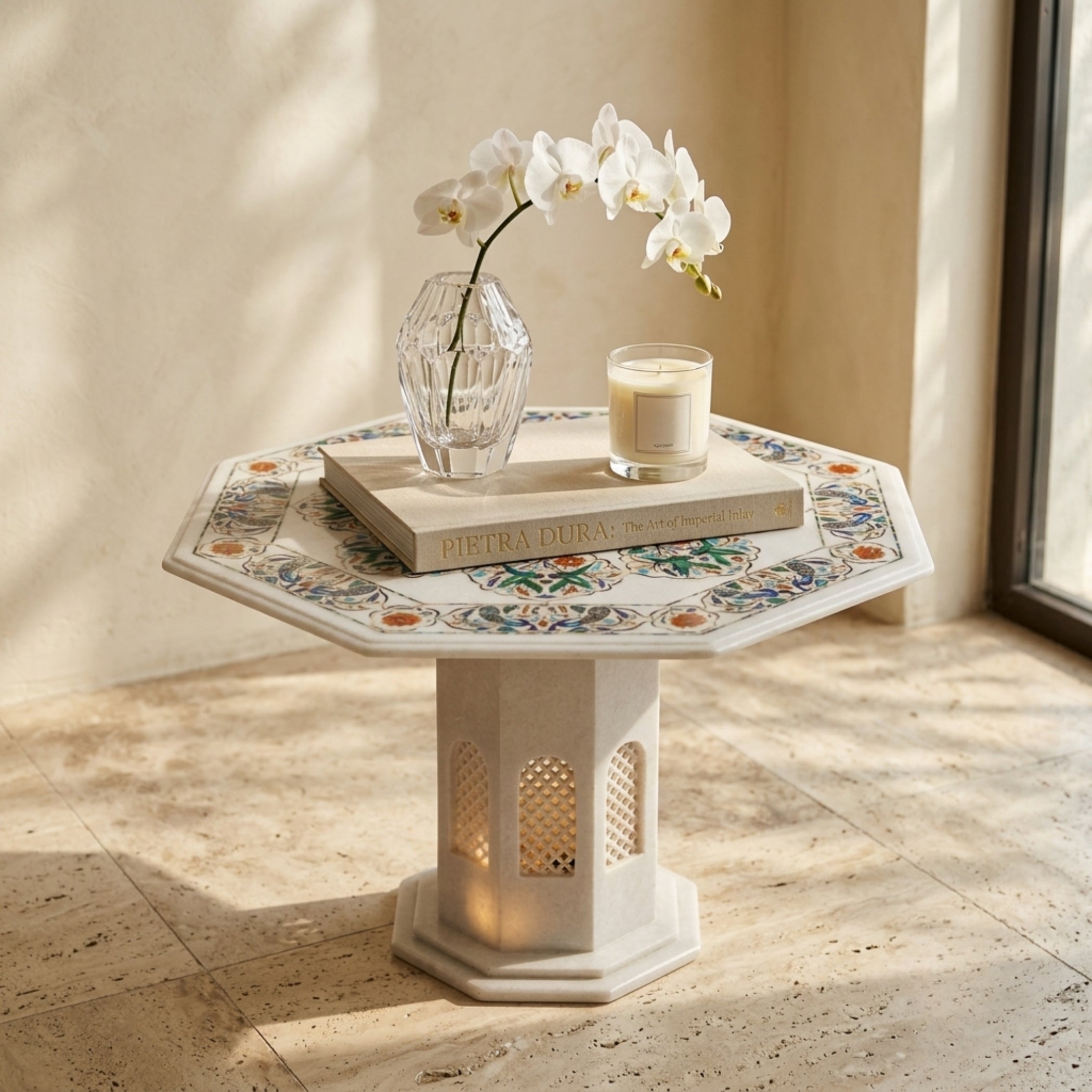 An octagonal side table with a mosaic tile top and a stone pedestal base, styled with a clear glass vase of white orchids, a book, and a candle in a sunlit interior corner.