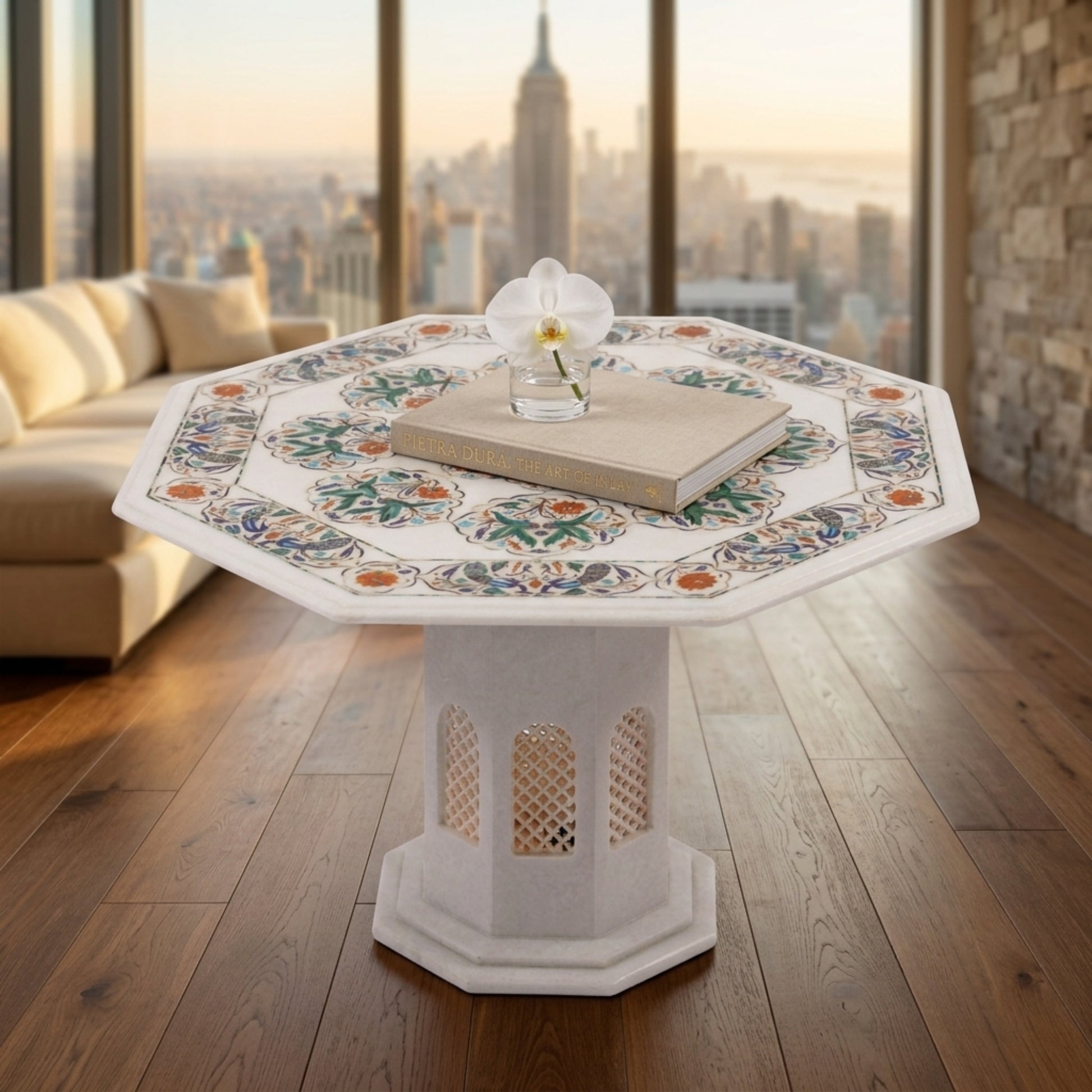 An octagonal white stone or concrete pedestal table with a colorful floral mosaic pattern on the tabletop, styled with a book and a small vase in a modern living room with a city skyline view.