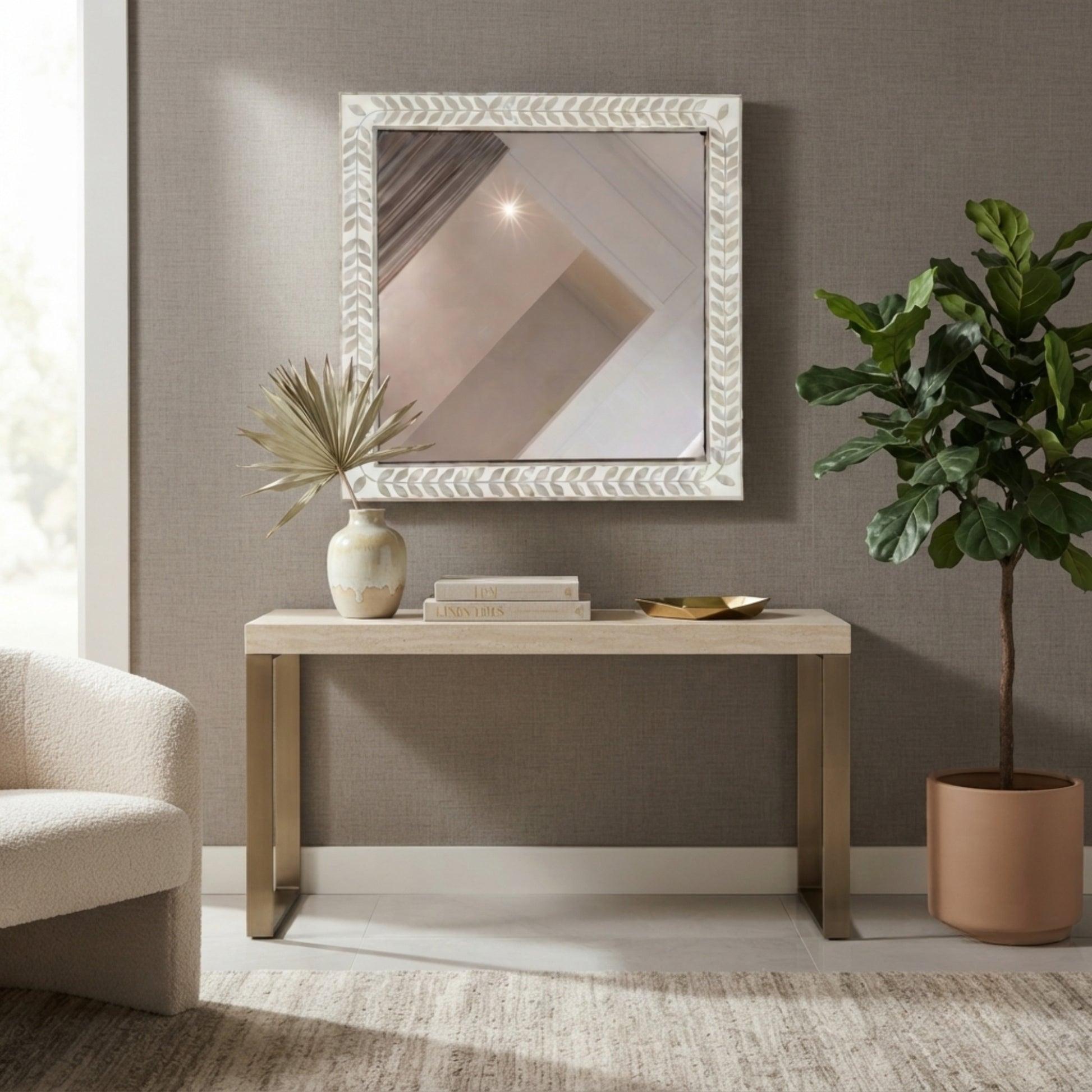 A square framed mirror with a textured white chevron border hangs on a taupe wall above a light wood console table. The scene is styled with a ceramic vase, books, a small bowl, a potted fiddle-leaf fig, and part of a cream armchair in a bright, modern room.