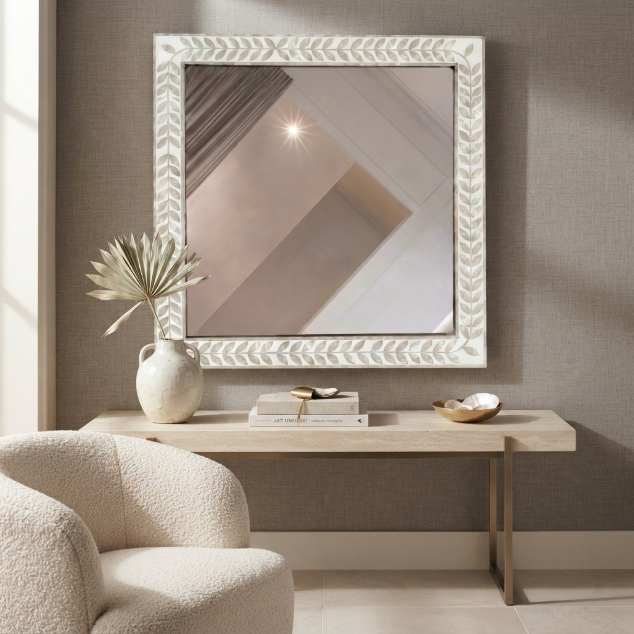 A square decorative mirror with a white carved leaf-patterned frame hangs on a textured taupe-gray wall above a light wood console table. A cream textured armchair is partially visible in the foreground, with a ceramic vase, books, and a small bowl on the table.
