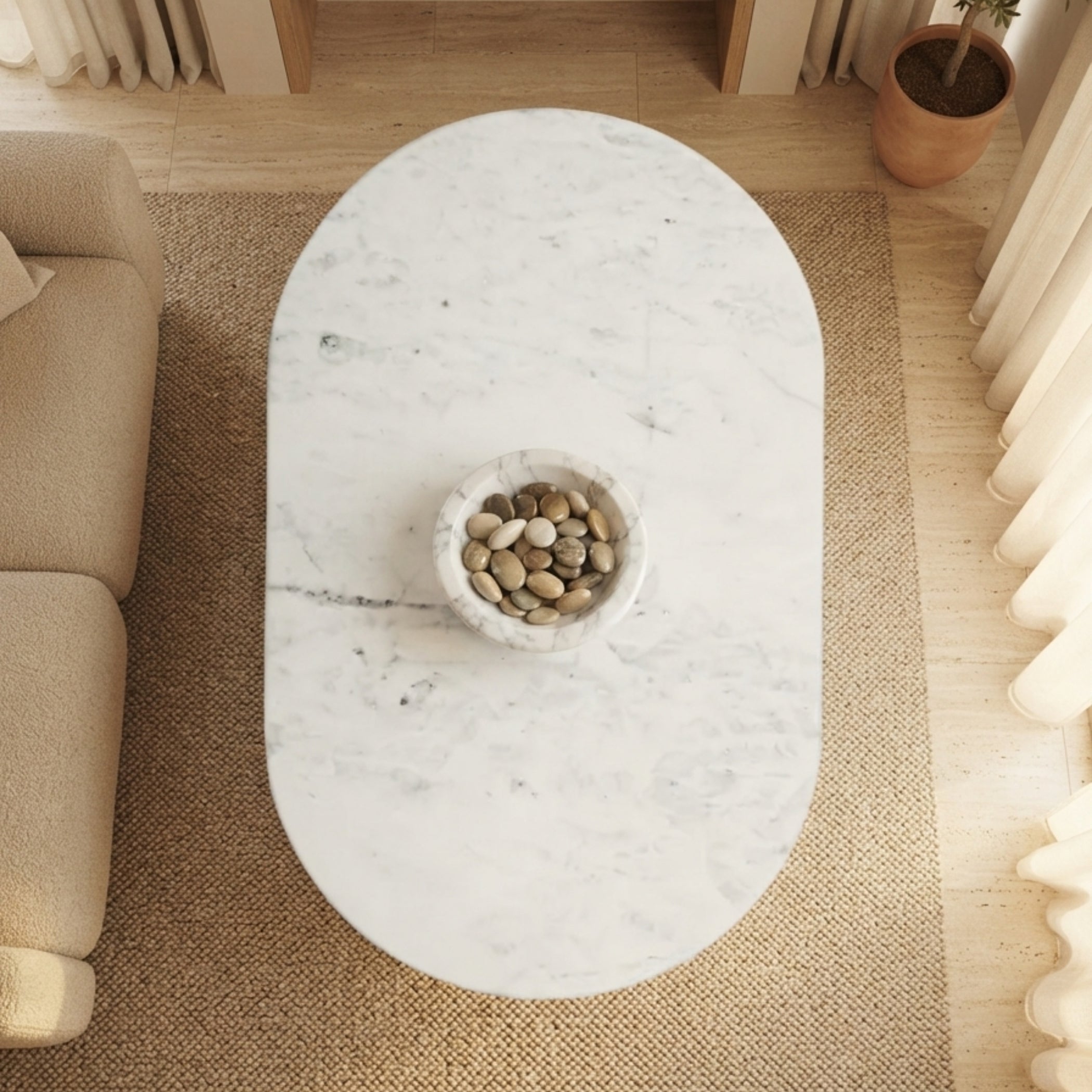An overhead view of an oval white marble coffee table with a central stone bowl, set on a woven jute rug in a modern living room.