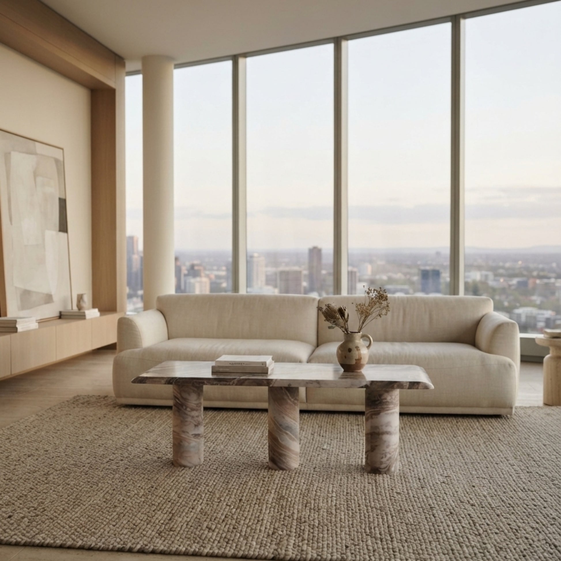 A modern living room featuring a cream-colored three-seater sofa, a marble-top coffee table with marble legs, a large textured beige rug, and floor-to-ceiling windows overlooking a city skyline.