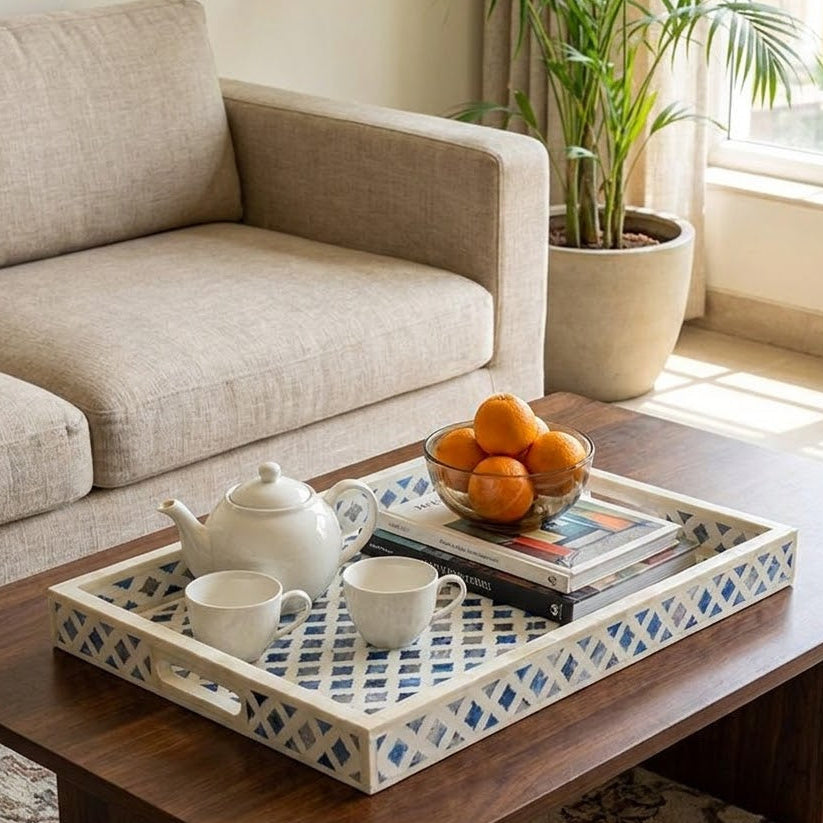 A rectangular decorative tray with a blue and white geometric lattice pattern and white handles, styled on a dark wood coffee table in a bright living room. The tray holds a white teapot, two white cups, a stack of books, and a glass bowl of oranges.