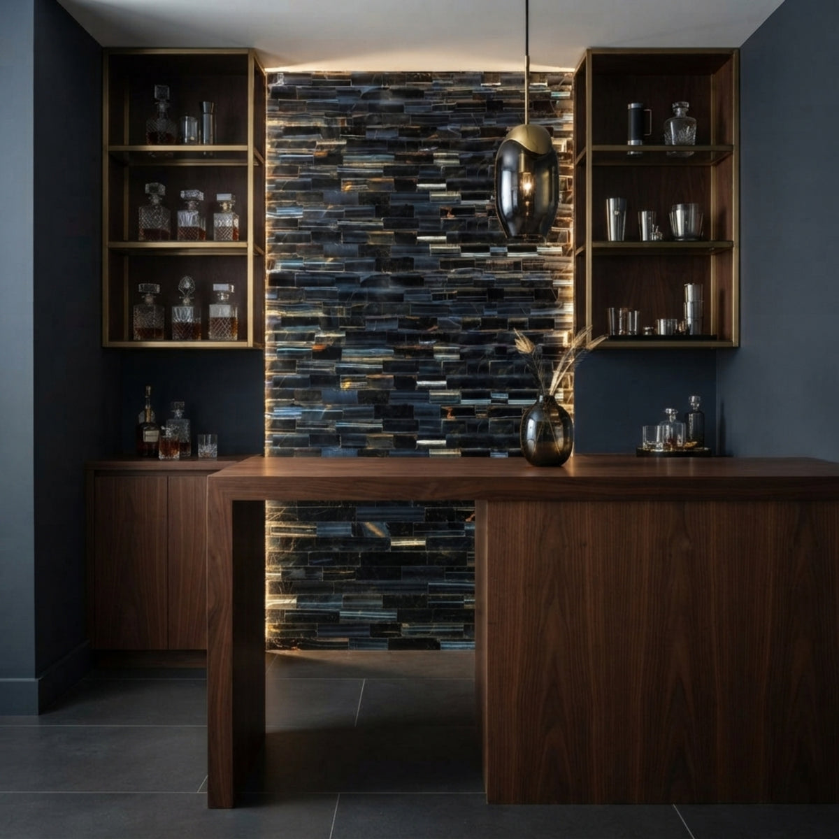 A modern bar or kitchen counter setup with a dark wood console table, matching side cabinets, open shelving filled with glassware, and a tall textured tile backsplash in blue, gray, and black tones. A dark pendant light hangs above.