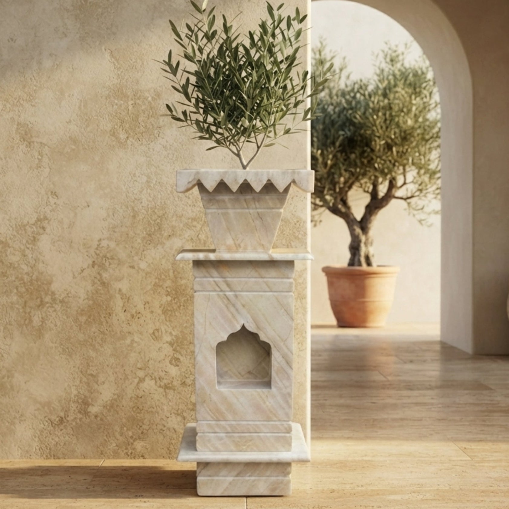 A tall white marble planter stand with a matching scalloped planter holding a green olive tree, set against a textured beige wall in an arched corridor with a terracotta pot in the background.