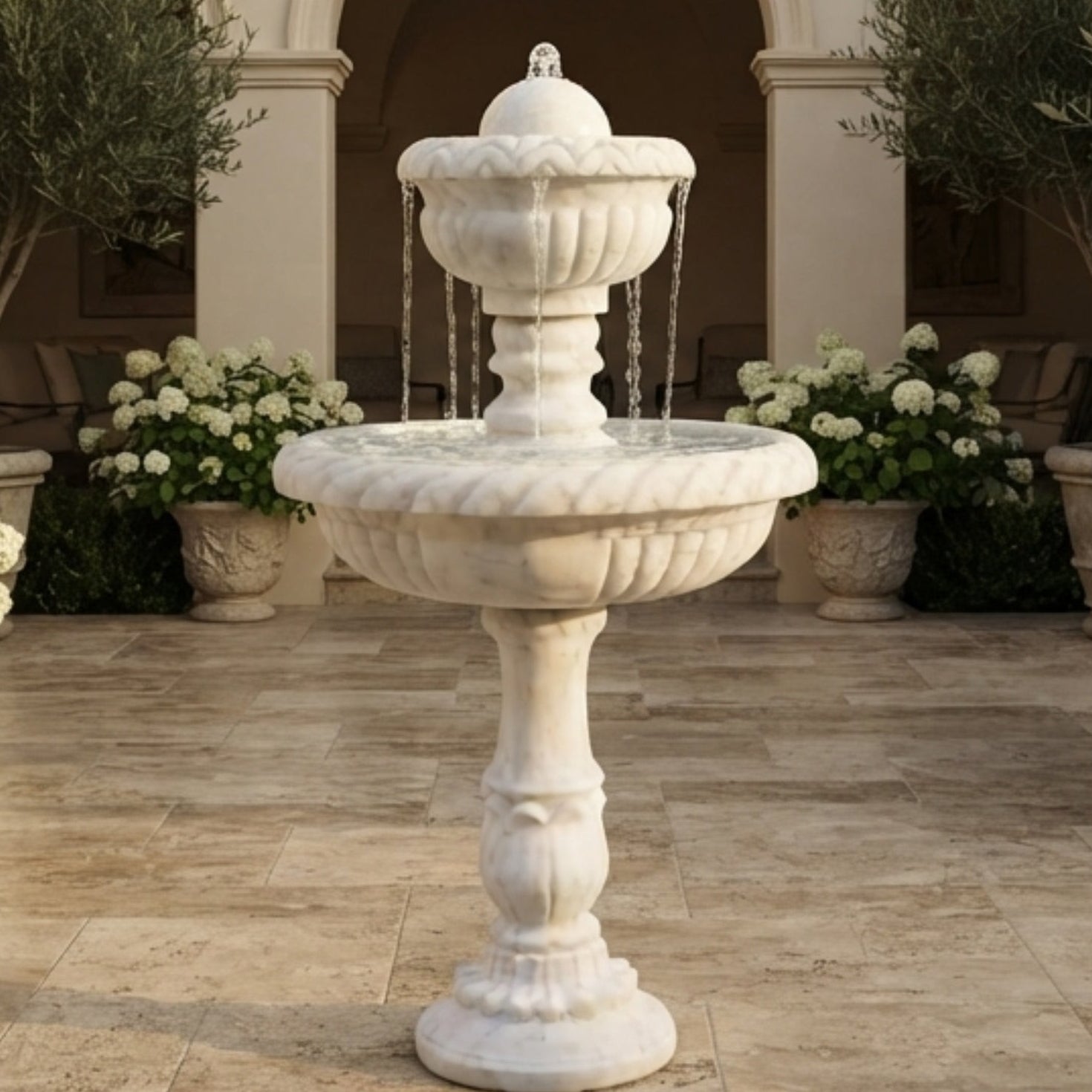 A white stone two-tiered fountain with water flowing from the top tier to the bottom basin, set in an outdoor courtyard with stone paving, potted olive trees, and white flower arrangements.