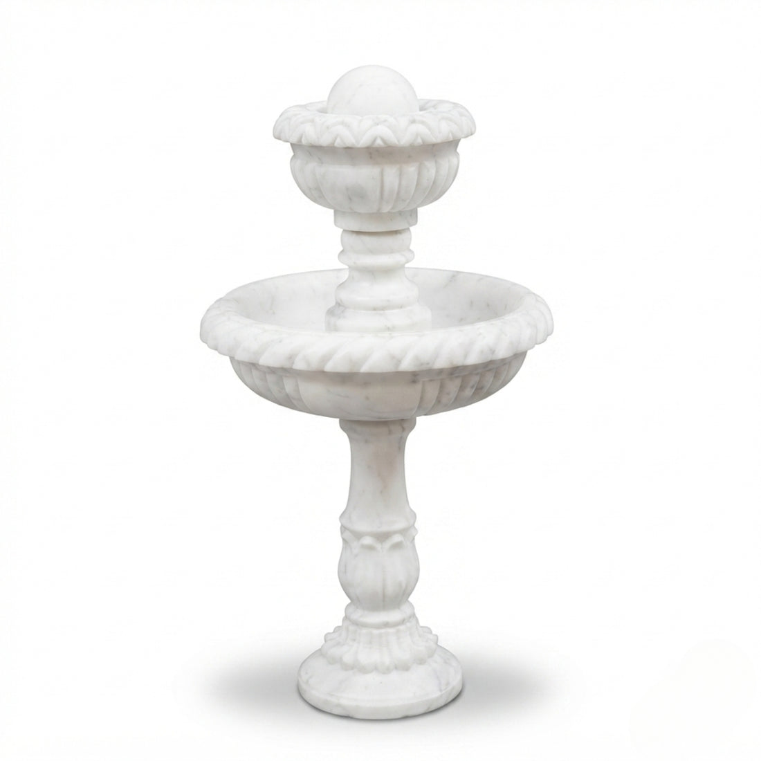 A white stone two-tiered fountain with water flowing from the top tier to the bottom basin, set in an outdoor courtyard with stone paving, potted olive trees, and white flower arrangements.