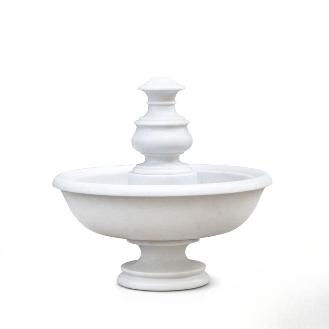 A tiered white stone fountain with water cascading down into a wide basin, set outdoors on a reflective dark surface with a modern house and landscaped garden in the background.