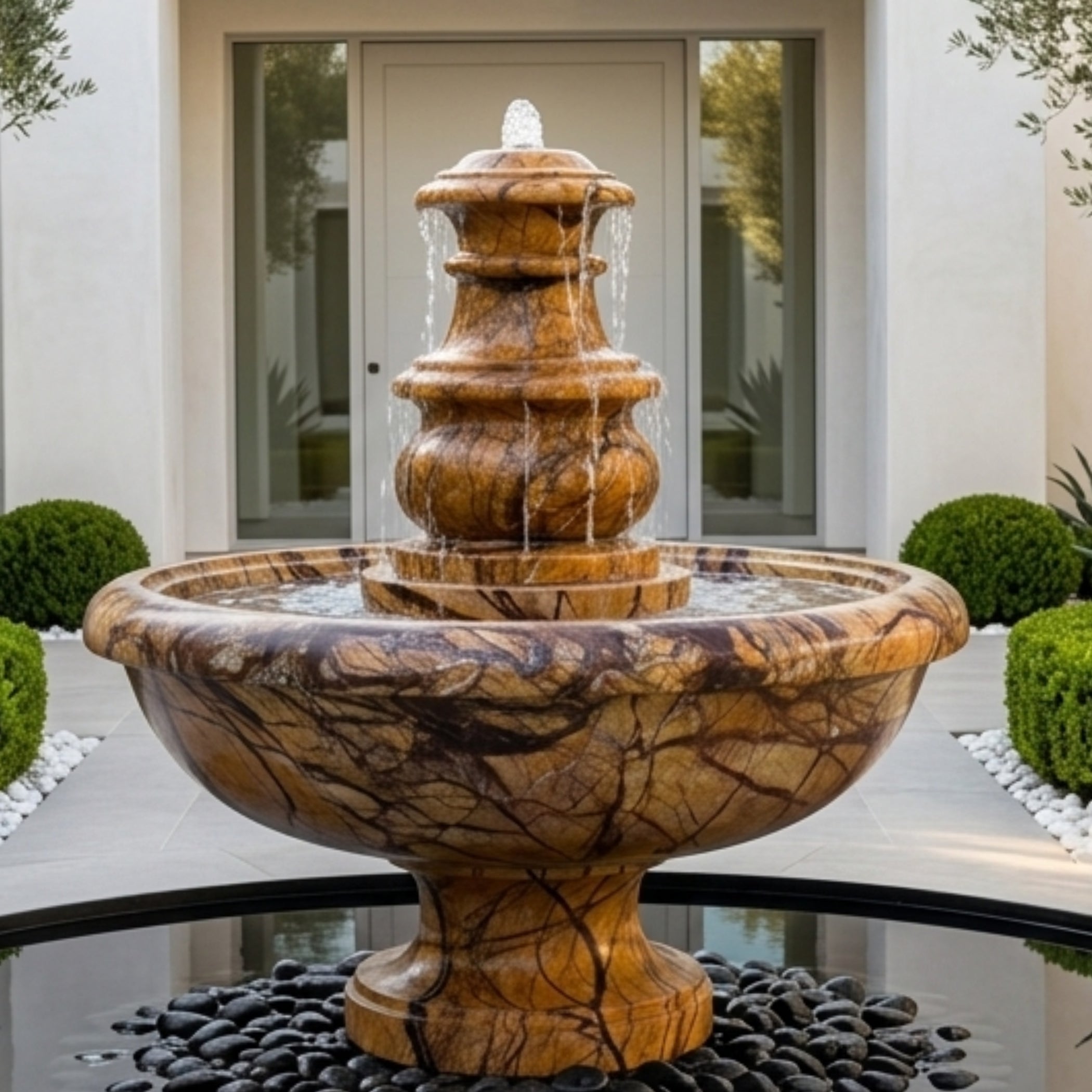 A tiered waterfall fountain made of polished brown and gold marbled stone, set outdoors in a garden courtyard with black river stones at the base.